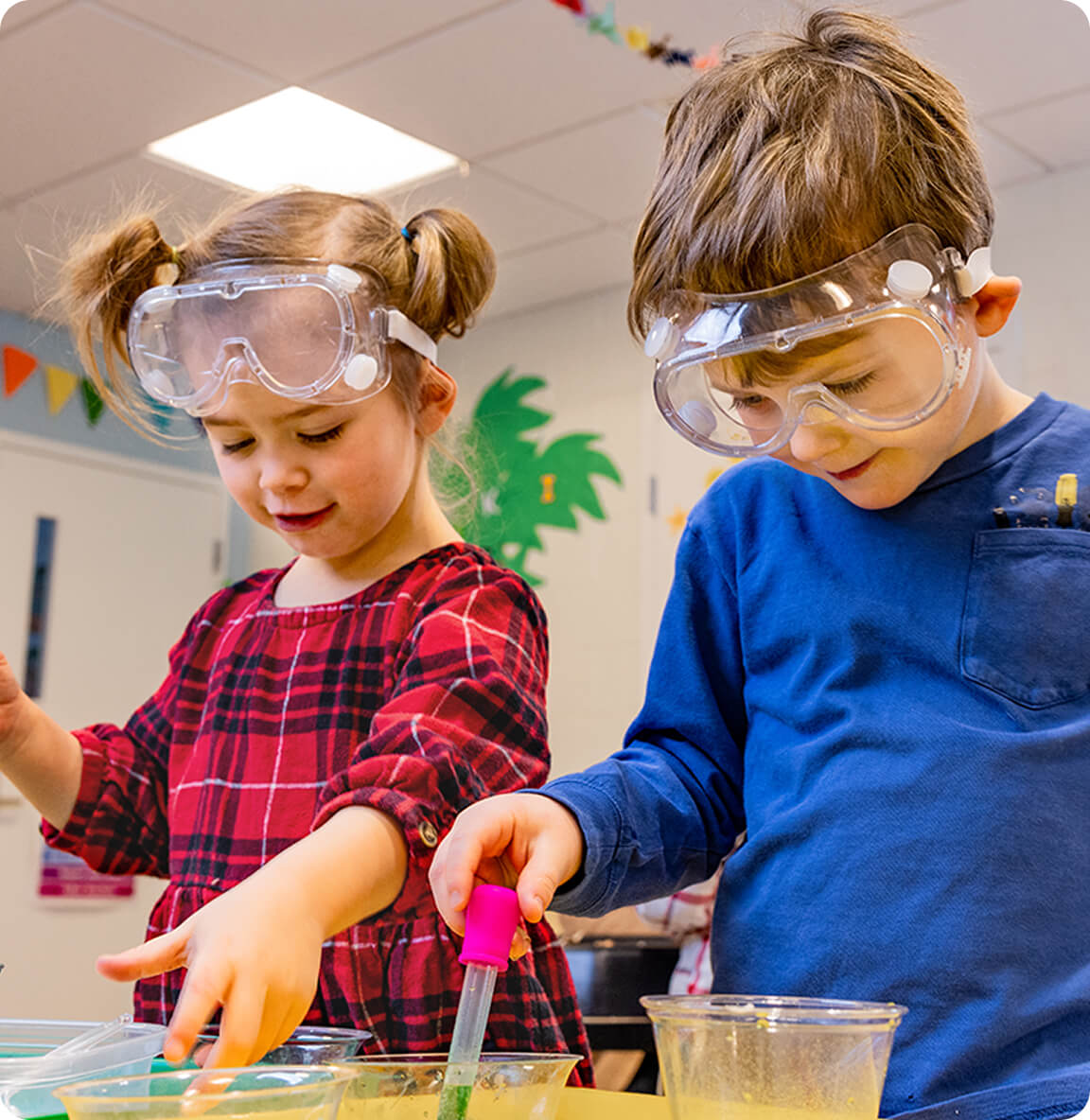 Two preschool children wearing safety goggles explore a classroom science activity with cups and droppers.