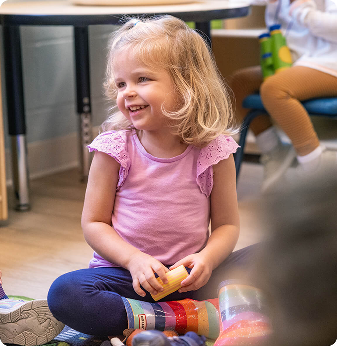 A smiling preschool girl sits on the classroom floor holding a toy block during group playtime.