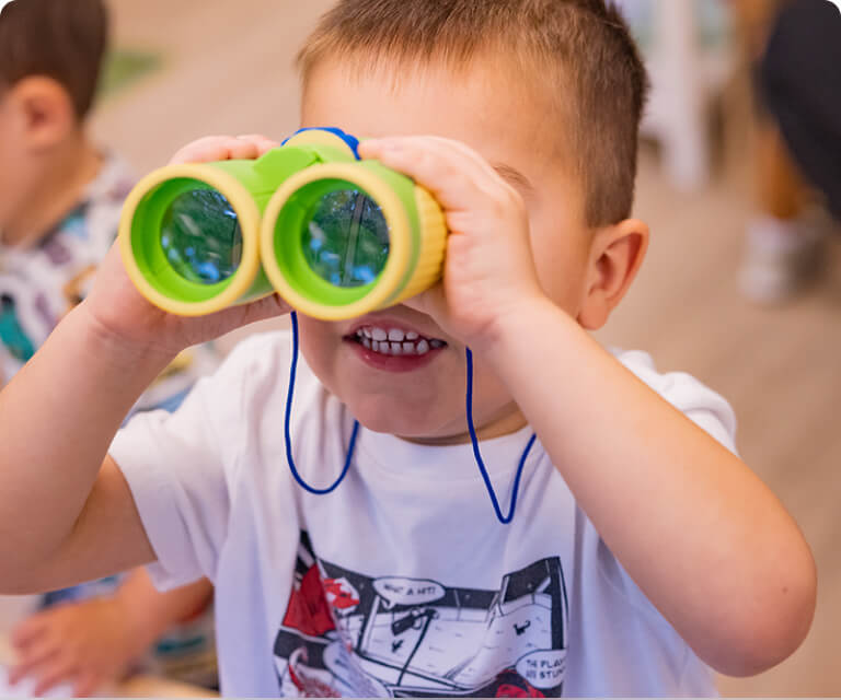 A preschool boy smiles while looking through bright green toy binoculars in the classroom.