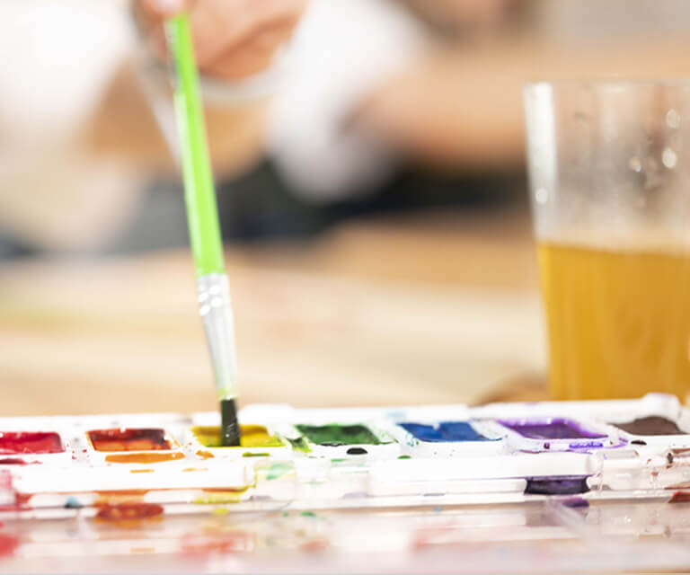 A child paints with watercolors, dipping a brush into a classroom paint tray beside a cup of water.